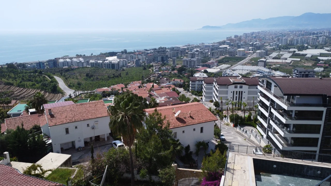 Zadivljujući pogled na more i grad iz nekretnine na obali. Ovaj panoramski eksterijer prikazuje živopisni turski obalni grad sa stambenim zgradama, bujnim zelenim brdima i svjetlucavim morem koje se proteže do udaljenih planina pod vedrim nebom.