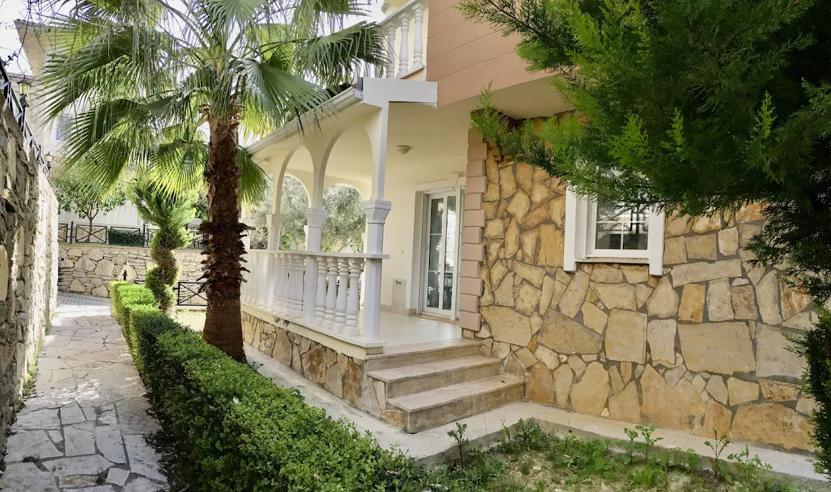Šarmantna ulazna vrata vile sa kamenom fasadom i stazom kroz vrt. Eksterijer šarmantne mediteranske vile sa rustičnom kamenom fasadom, lučnim trijemom, bijelom balustradom i popločanom stazom kroz vrt sa bujnim palmama.