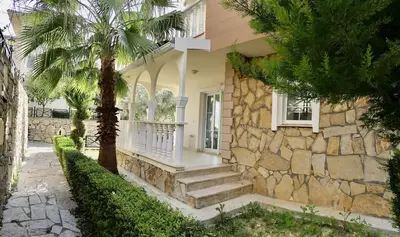 Charming Villa Entrance with Stone Facade and Garden Path. Exterior of a charming Mediterranean villa featuring a rustic stone facade, arched porch, white balustrade, and a paved garden path with lush palm trees.