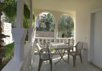 Charming Balcony with Garden View and Arched Architecture. A charming, covered balcony featuring a tiled floor, white arched pillars, and a classic railing. It includes a plastic table and chairs, offering a serene view of a green garden with trees.