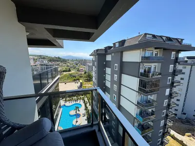 Moderni balkon s bazenom i panoramskim pogledom na grad. Moderni balkon stana sa staklenom ogradom, garniturom za sjedenje i zadivljujućim pogledom na veliki vanjski bazen, susjedne zgrade i udaljene planine.