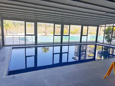 Moderan zatvoreni bazen s panoramskim prozorima. Moderan, dobro održavan zatvoreni bazen s tamnoplavim pločicama. Veliki panoramski prozori nude pogled na vanjski bazen i bujno zelenilo, osiguravajući obilje prirodnog svjetla.