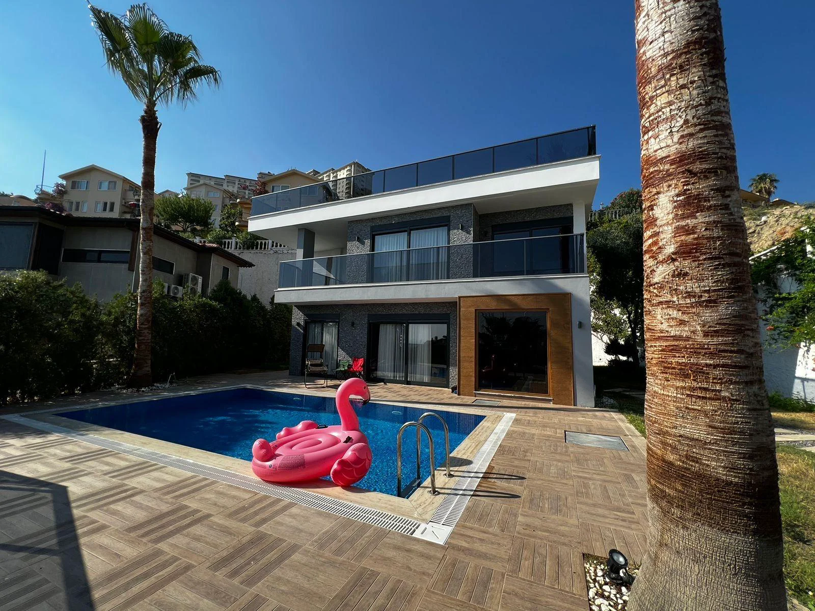 Luksuzna moderna vila s privatnim bazenom. Eksterijer moderne vile koji prikazuje privatni bazen s ružičastim flamingom na napuhavanje, elegantnu popločanu terasu i staklene balkone pod sunčanim nebom s palmama.