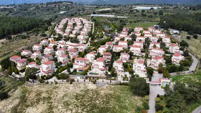 Aerial View of Luxury Hillside Villa Complex. Aerial view of a modern residential complex with numerous two-story villas, red roofs, and light facades. Features include green landscaping, a swimming pool, and natural hillside surroundings.