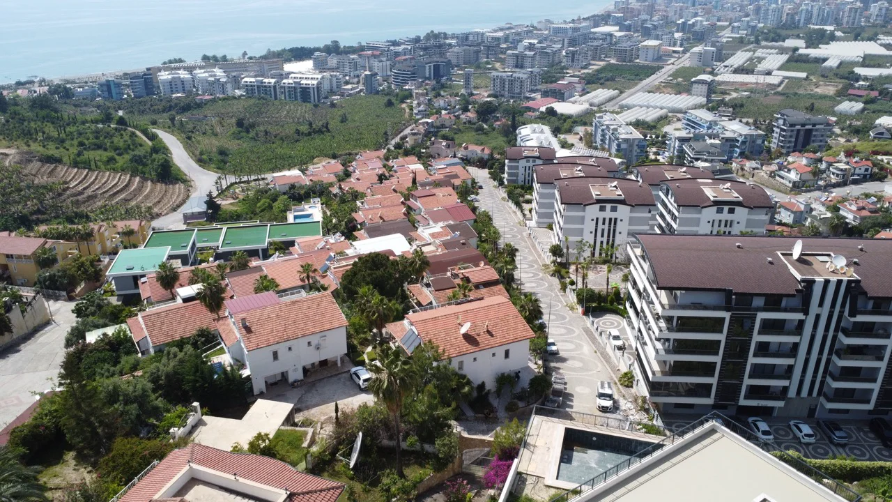 Pogled iz zraka na stambenu četvrt, turska obala s pogledom na more. Opsežna fotografija iz zraka turskog obalnog stambenog područja, s tradicionalnim vilama s crvenim krovovima i modernim stambenim kompleksima. Slika uključuje zelena terasasta brda, gradske ceste i svjetlucavo Sredozemno more u daljini.
