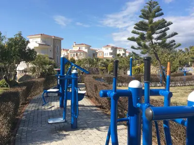 Outdoor Fitness Equipment in Residential Development. An outdoor fitness area features modern blue exercise equipment on a paved pathway, surrounded by green bushes and trees, with residential buildings visible under a clear blue sky.