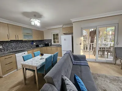 Open Plan Kitchen and Living Room with Terrace Access. A modern open plan kitchen and living room. It features wooden cabinetry, mosaic backsplash, a dining area, and a comfortable grey sofa. A sliding glass door opens to an outdoor patio.