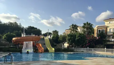 Resort-Style Outdoor Pool with Water Slides. An inviting outdoor swimming pool with blue water and two large water slides. People are seen enjoying the water. The pool area is surrounded by lush green trees, palm trees, and residential buildings under a clear sky.