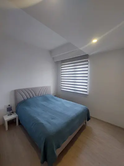 Moderna spavaća soba sa svijetlim drvenim podom i žaluzinama. Moderna spavaća soba sa svijetlim drvenim laminatom i bijelim zidovima. Sadrži bračni krevet sa sivim teksturiranim uzglavljem, plavim prekrivačem i noćnim ormarićem s lampom. Prozor s horizontalnim žaluzinama pruža privatnost.