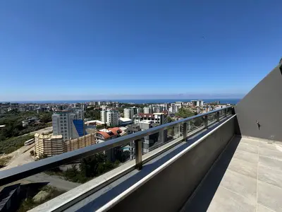 Zapanjujući panoramski pogled na more i grad s balkona. Moderni balkon sa sivim popločanim podom i elegantnom stakleno-metalnom ogradom, pruža zapanjujući panoramski pogled na grad, bujne zelene površine i udaljeno plavo more pod vedrim nebom.