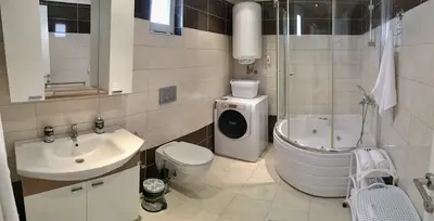 Modern Bathroom with Jacuzzi, Washing Machine and Vanity. A modern bathroom featuring a vanity sink, wall-mounted toilet, washing machine, and a corner shower with a built-in jacuzzi bathtub. Tiled in light beige and brown.