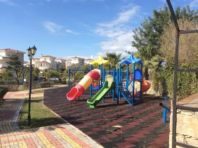 Vibrant Outdoor Children's Playground in Residential Community. A vibrant outdoor children's playground featuring colorful slides and climbing frames on safety rubber flooring. Set in a sunny residential community, surrounded by green landscaping and modern villas, perfect for families.