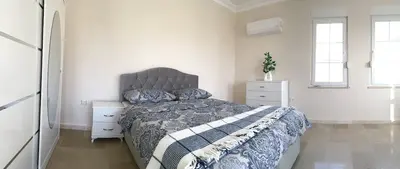 Bright Modern Bedroom with Comfortable Bed and Storage. A modern bedroom featuring a comfortable bed with a grey upholstered headboard, patterned bedding, tiled flooring, a white wardrobe, bright windows, and an air conditioning unit.