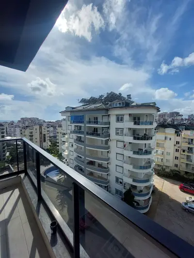 Prostrani balkon sa pogledom na grad i more u daljini. Moderni balkon sa popločanim podom i elegantnom staklenom ogradom, koji nudi panoramski pogled na užurbani grad, mali bazen i more u daljini pod vedrim, djelimično oblačnim nebom.