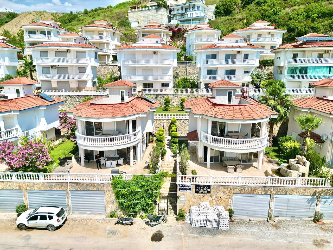 Kompleks vila na brdu s balkonima i uređenim vrtovima. Pogled iz zraka na moderni stambeni kompleks na brdu u Turskoj, koji prikazuje višespratne bijele vile s crvenim krovovima, privatnim balkonima i bujnim zelenim pejzažom.