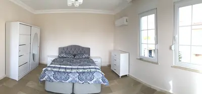 Spacious Bedroom with Tiled Floor and Modern Furniture. Modern bedroom with light tiled flooring. Features a double bed with grey upholstered headboard, white wardrobe with mirror, dresser, two large windows, and an AC unit.