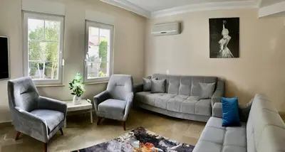 Modern Living Room with Garden Views and Comfortable Seating. A bright, modern living room with light marble flooring and large windows showcasing a green garden view. Features comfortable grey sofas, an air conditioner, and a decorative floral rug.
