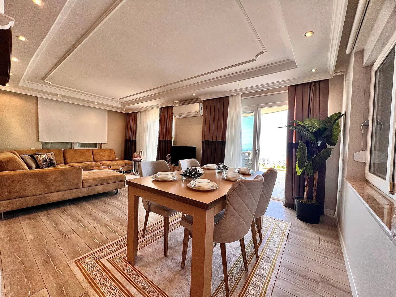 Savremeni dnevni boravak sa elegantnom trpezarijom i pogledom na more. Moderni dnevni boravak i trpezarija sa drvenim podovima i ugradnom rasvjetom. Sadrži udobnu sofu u obliku slova L, elegantnu trpezarijsku garnituru i izlaz na balkon sa pogledom na more.