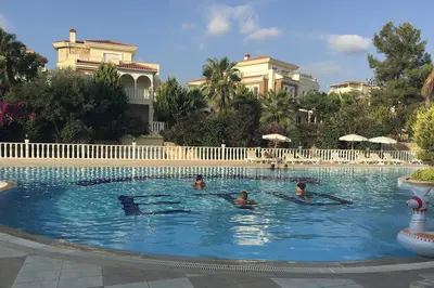 Spacious Outdoor Pool in Residential Complex with Villas. A vibrant outdoor swimming pool area in a residential complex on a sunny day. Features a large pool with people, sun loungers, umbrellas, and traditional villas surrounded by lush gardens.