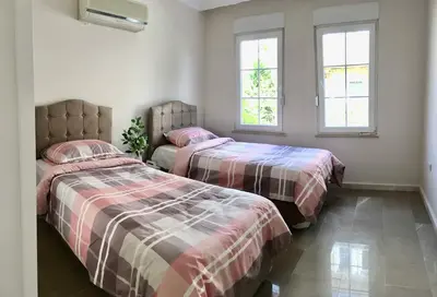 Bright Twin Bedroom with Air Conditioning and Tiled Floor. A bright, modern bedroom featuring two single beds with tufted headboards, plaid bedding, and grey tiled flooring. Two windows provide natural light; a wall-mounted air conditioner is visible.