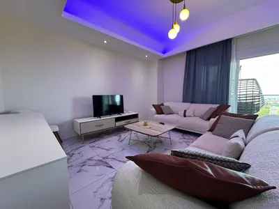 Savremeni dnevni boravak sa ambijentalnim LED osvjetljenjem. Moderan dnevni boravak sa podovima od pločica nalik mermeru, stilskim L-sofe, televizorom ravnog ekrana i jedinstvenim ljubičastim LED plafonskim osvjetljenjem. Balkon sa vanjskim sjedenjem je vidljiv kroz prozor.