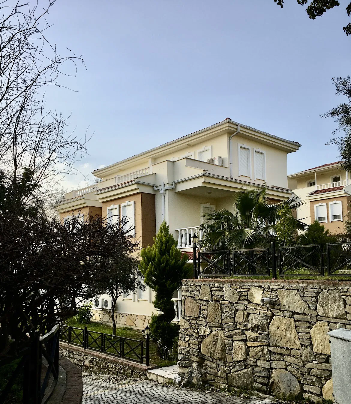 Moderna vila sa vrtom i kamenim zidom. Eksterijer moderne, krem boje, višespratne vile sa nekoliko prozora i malih balkona. Ima njegovani vrt, čvrsti potporni zid od prirodnog kamena i popločani ulazni put.