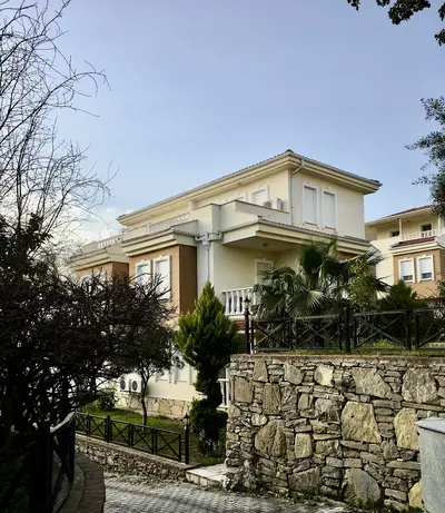 Modern Villa Exterior with Garden and Stone Wall. The exterior of a modern, cream-colored multi-story villa with several windows and small balconies. Features a well-maintained garden, a sturdy natural stone retaining wall, and a paved entrance path.