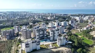 Panoramski pogled na stambeni kompleks s pogledom na more, iz zraka. Prostrani pogled iz zraka na moderan stambeni kompleks s više zgrada, zelenim površinama i vanjskim bazenom, s udaljenim gradom i horizontom plavog mora pod vedrim nebom.