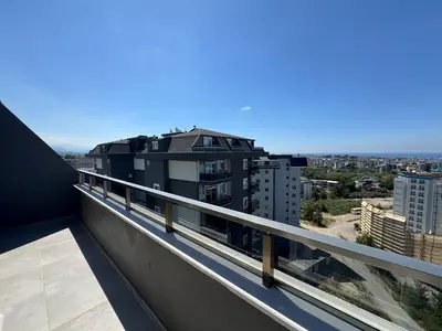 Sunčani balkon sa zadivljujućim gradskim pejzažom i pogledom na ocean. Moderan balkon sa svijetlo sivim pločicama i elegantnom stakleno-čeličnom ogradom, pruža neometan panoramski pogled na gradski horizont, bujne zelene površine i pjenušavo more pod vedrim plavim nebom.
