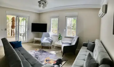 Bright Modern Living Room with Balcony and Garden View. Bright, modern living room with light flooring, grey sofas, armchairs, and a wall-mounted TV. Features air conditioning, a patterned rug, and balcony access with garden views.