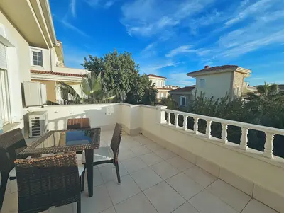 Spacious Terrace with Outdoor Dining and Neighborhood View. A bright, spacious outdoor terrace with light-colored floor tiles and a classic white balustrade. It features a woven outdoor dining set for four, offering views of neighboring villas and lush green trees under a clear sky.