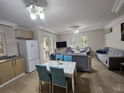Modern Open-Plan Living Area with Dining and Kitchenette. An inviting open-plan living and dining area with modern grey sofas and a dining table. Features tiled flooring, ceiling lights, air conditioning, and a small kitchenette with outdoor access.