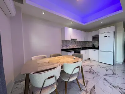 Moderni prostor za objedovanje u kuhinji sa stilskim osvjetljenjem. Moderna kuhinja i trpezarija sa elegantnim bijelim ormarićima, crnim pultovima i sjajnim podom nalik mermeru. Sadrži moderan trpezarijski set, ljubičasto ambijentalno osvjetljenje i klima uređaj.
