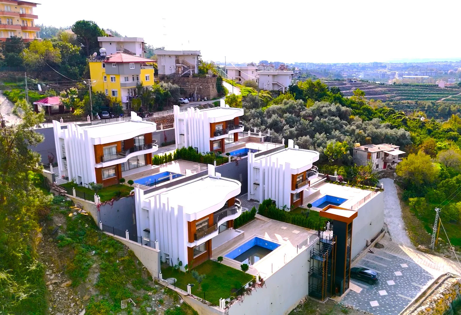 Luxury Hillside Villas with Private Swimming Pools. Aerial view of a modern residential complex featuring multiple contemporary villas. Each home includes a private swimming pool, terraced gardens, and stunning hillside views.