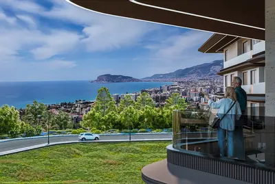 Panoramic Sea and City Views from a Modern Balcony. A couple enjoys the panoramic Mediterranean Sea and Alanya city views from a modern apartment balcony. The balcony features a sleek glass railing, offering an unobstructed, luxurious vista.