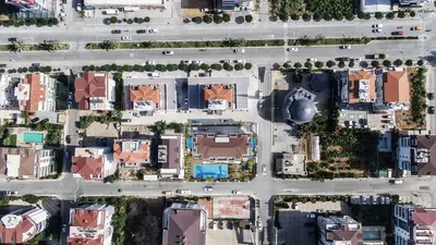 Aerial View of Residential Area with Swimming Pool. An aerial drone shot showcasing a vibrant residential neighborhood with multiple red-roofed houses, roads with cars, green spaces, and a prominent property featuring a large, inviting blue swimming pool.