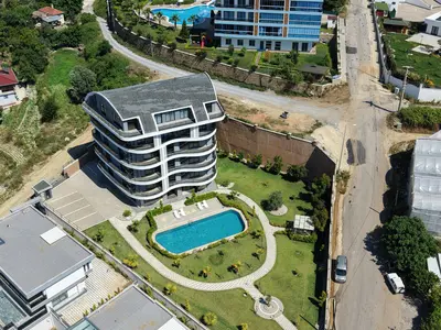Modern Apartment Building with Private Pool and Lush Garden. Aerial view of a modern, curved apartment building. Features a private outdoor swimming pool, lush green garden with pathways, lounge chairs, and an adjacent parking area, all well-maintained.