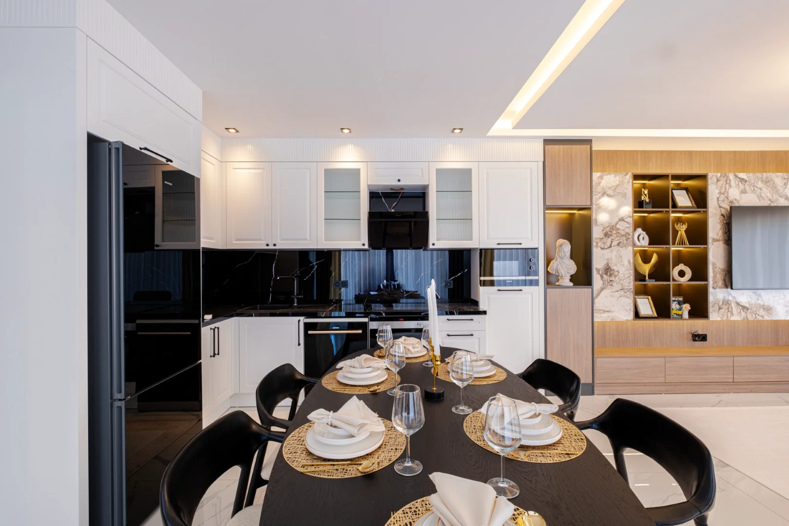 Modern Open-Concept Kitchen and Dining Area. A modern open-concept kitchen and dining area, featuring sleek white cabinetry, black marble backsplash, and integrated appliances. A dark oval dining table is elegantly set in the foreground, illuminated by contemporary lighting.
