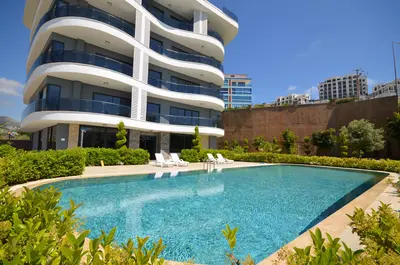 Modern Apartment Complex with Refreshing Outdoor Swimming Pool. View of a modern apartment complex exterior featuring a large, clean outdoor swimming pool in the foreground, surrounded by lush green landscaping and white sun loungers on a bright sunny day.