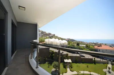 Modern Balcony with Panoramic Sea and Garden Views. A modern balcony featuring grey walls, tiled flooring, and a sleek glass-and-metal railing. It offers panoramic views of a lush green garden, distant greenhouses, and the clear blue Mediterranean Sea.