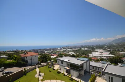 Panoramic Coastal City and Mountain Vista. A breathtaking outdoor view featuring the bright blue Mediterranean Sea, a sprawling coastal city, green hills, and distant mountains under a clear sky. Includes modern buildings and a green garden.