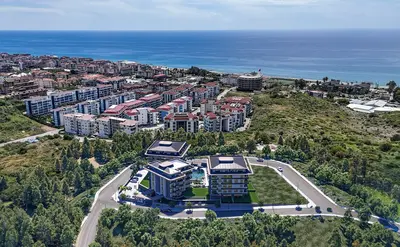 New Coastal Residential Project Overlooking the Mediterranean Sea. Aerial view of a new coastal residential complex featuring modern buildings, a swimming pool, and green areas. It boasts panoramic views of the Mediterranean Sea and the nearby town.