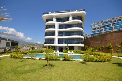 Modern Architectural Building with Pool and Landscaped Grounds. Modern residential building exterior with a private swimming pool, green lawn, and landscaped garden. Clear blue sky and distant mountains complete this well-maintained property view.