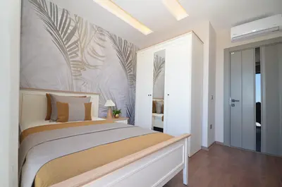 Modern Bedroom with Stylish Palm Leaf Wallpaper and AC. A modern bedroom featuring light wood flooring, stylish palm leaf wallpaper, a comfortable bed, and a large white wardrobe with mirrored doors. Includes recessed lighting and an air conditioning unit.