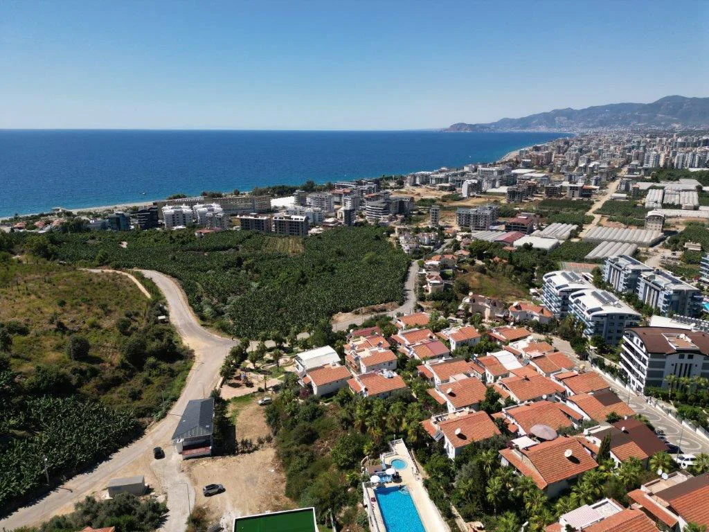 Panoramisk hav- og byudsigt over Alanyas kystlinje. En omfattende luftudsigt, der viser en tyrkisk kystby med det dybblå Middelhav, sandstrand, adskillige bygninger, grønne bananplantager og en privat swimmingpool under en klar himmel.