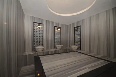 Luxurious Marble Turkish Bath with Traditional Design. Luxurious Turkish bath with elegant gray and white marble walls and surfaces. Features three basins, ornate gold faucets, tiled insets, and a large central marble slab for relaxation.