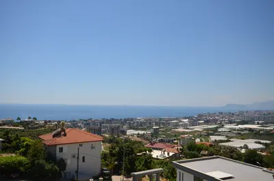 Panoramic Sea and City View from Coastal Property in Turkey. A wide exterior shot revealing a stunning sea and city panorama under a clear blue sky. The vibrant view includes distant mountains, urban buildings, and lush green landscape.
