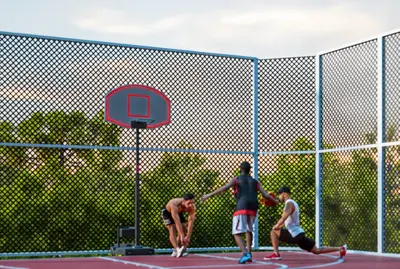 Modern Basketball Court for Active Lifestyle. An inviting outdoor basketball court with a red playing surface and a portable hoop. Three men are visible, one stretching and two playing. Lush green trees and a fence surround the modern sports facility.