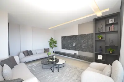 Stylish Modern Living Room with Elegant Decor and Entertainment Unit. A modern, elegantly decorated living room featuring comfortable grey sofas, a large marble-effect TV wall, recessed lighting, and stylish built-in shelves with decor. It's a bright, welcoming space.