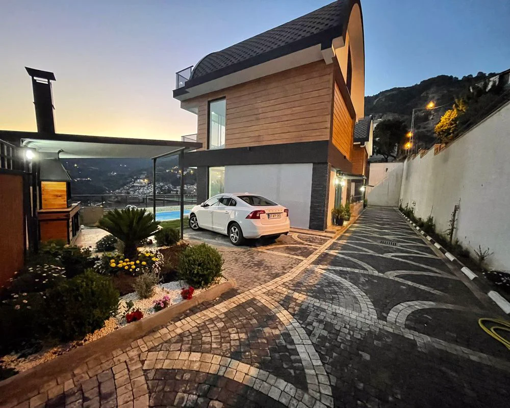 Moderne villa-eksteriør med pool, have og panoramaudsigt. Moderne villa-eksteriør i skumringen med unik arkitektur, en brolagt indkørsel og anlagt have. Indeholder et udekøkken, privat pool og en fantastisk panoramaudsigt over by og bjerge.